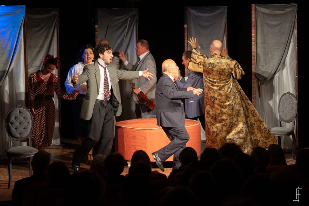 Attori in scena durante la rappresentazione teatrale de "Il medico dei pazzi" con un gruppo di persone in un contesto scenico teatrale.