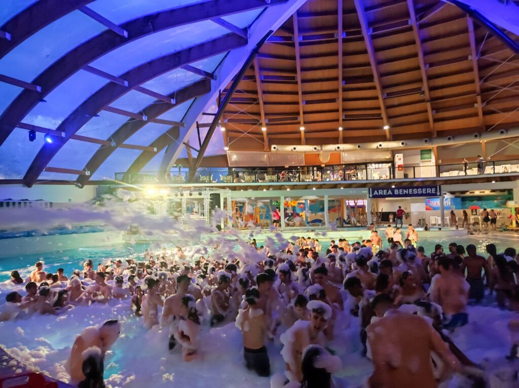 Foto di persone in piscina durante una festa con schiuma in un parco acquatico coperto.