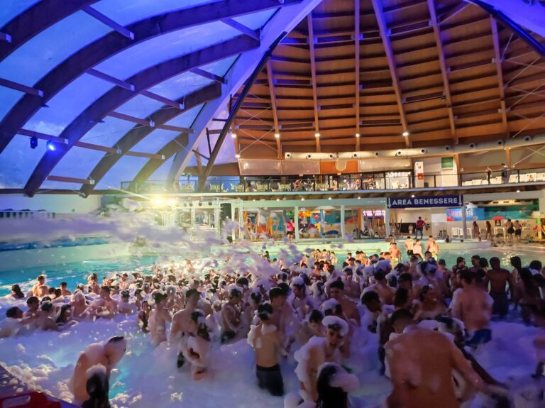 Foto di persone in piscina durante una festa con schiuma in un parco acquatico coperto.
