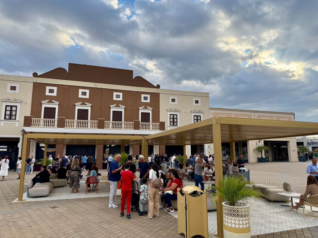 Persone radunate in una piazza esterna davanti a un edificio, con un cielo nuvoloso in serata.