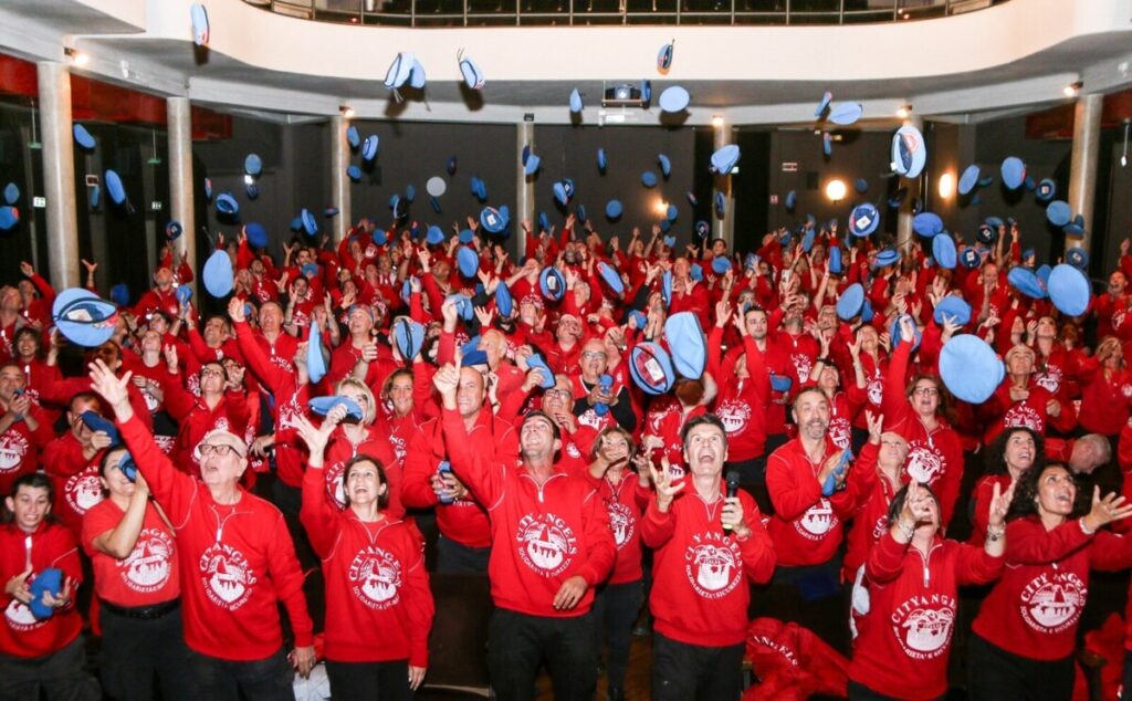 Volontari dei City Angels durante un raduno, indossano maglie rosse e lanciano in aria cappellini blu in una sala interna.